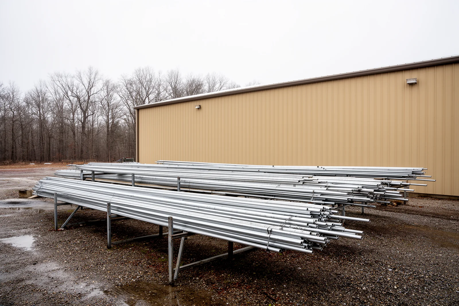 Long aluminum tubing stacked on racks outside a low industrial building on a flat eastern North Carolina site under an overcast sky